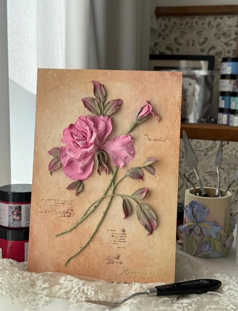 May include: A textured artwork featuring a pink rose with green stems and leaves against a beige background. The rose is in full bloom with detailed petals. The artwork has a vintage aesthetic, with script-like text and a signature at the bottom.