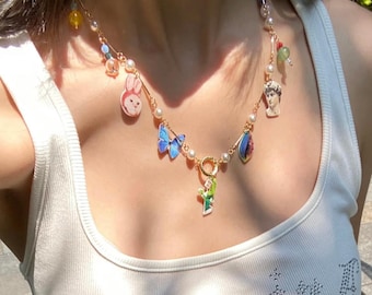 May include: A gold-toned necklace featuring a variety of charms, including a blue butterfly, a bunny, a small statue, and a green figure. The necklace also includes pearls and colorful beads, creating a whimsical and artistic design.