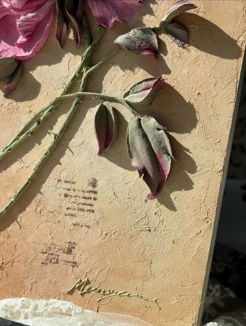 May include: A textured, rectangular artwork featuring a detailed relief of a pink rose with green stem and leaves. The background is a light tan color with faded text and a signature. The artwork is a unique piece of floral art.