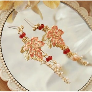 May include: A pair of gold-toned hairpins featuring a maple leaf design with red and peach accents. The hairpins have dangling chains with small pearls and red beads, resting on a reflective surface.