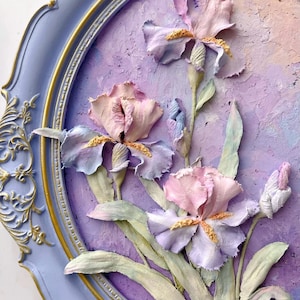 May include: A framed floral artwork featuring three-dimensional iris flowers in shades of lavender, pink, and white. The flowers are set against a textured purple background within a light blue oval frame with gold accents. The artwork has a vintage aesthetic.