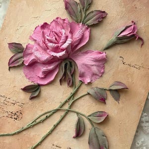 May include: A pink rose sculpture with green stems and leaves, set against a textured, tan background. The rose is in full bloom with detailed petals, and a bud is also visible. The artwork has a vintage aesthetic.