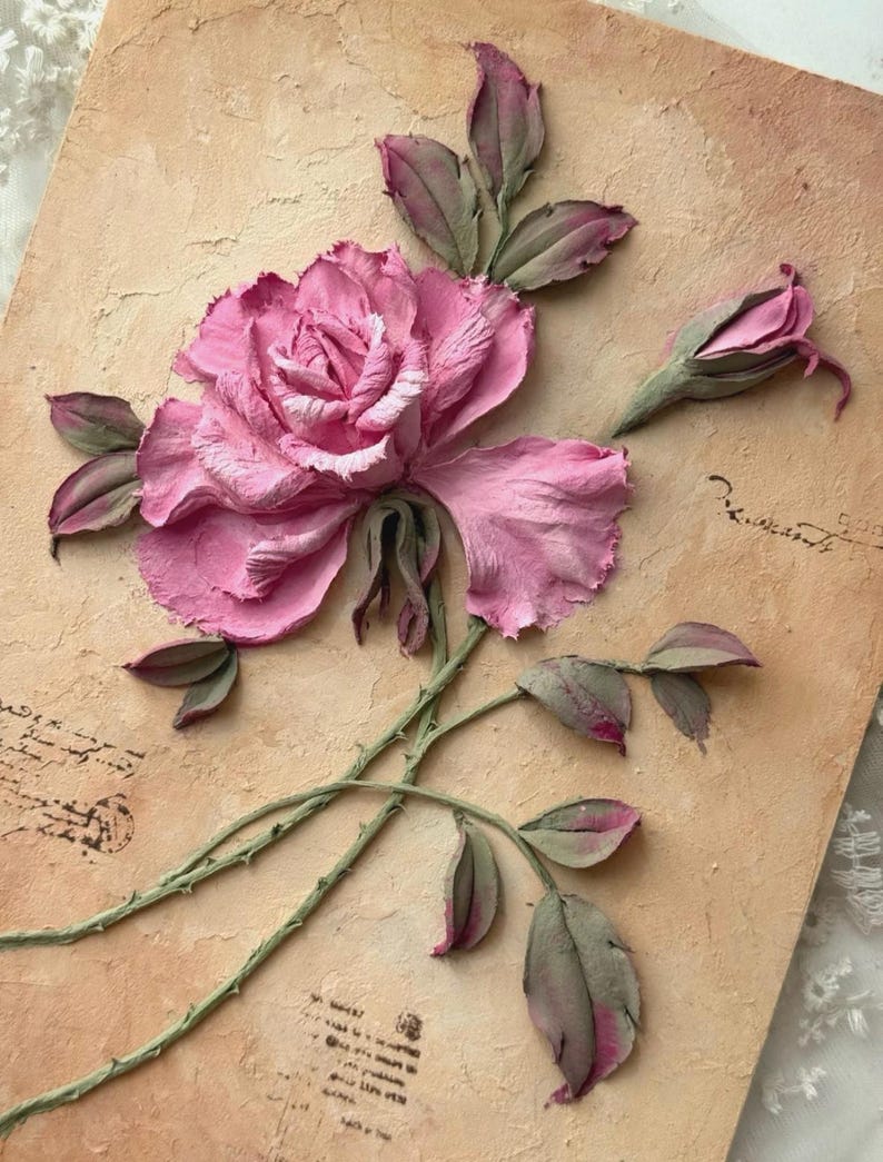 May include: A pink rose sculpture with green stems and leaves, set against a textured, tan background. The rose is in full bloom with detailed petals, and a bud is also visible. The artwork has a vintage aesthetic.