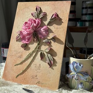 May include: A textured artwork featuring a pink rose with green stems and leaves. The rose is the focal point, set against a beige background with script-like text. A palette knife rests in the foreground.