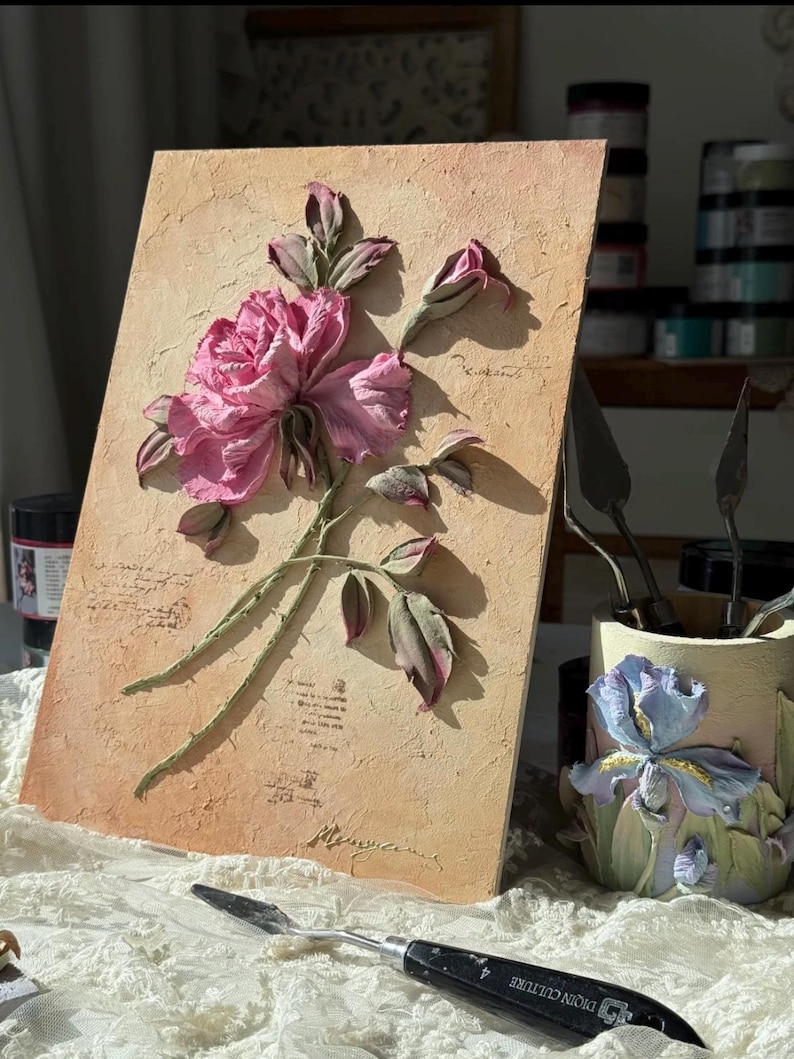 May include: A textured artwork featuring a pink rose with green stems and leaves. The rose is the focal point, set against a beige background with script-like text. A palette knife rests in the foreground.