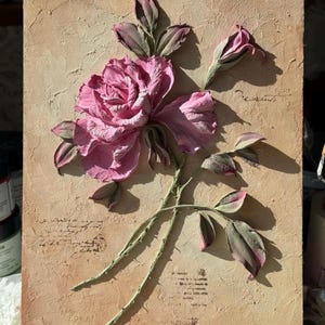 May include: A textured, rectangular artwork featuring a raised relief of a pink rose with green stems and leaves. The background is a light brown with faded script. The rose is in full bloom with detailed petals and a bud.