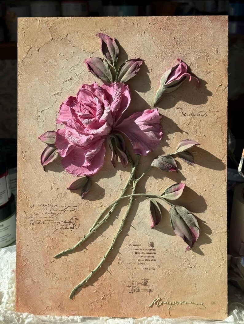 May include: A textured, rectangular artwork featuring a raised relief of a pink rose with green stems and leaves. The background is a light brown with faded script. The rose is in full bloom with detailed petals and a bud.