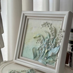May include: A framed artwork with a 3D relief of lily of the valley flowers in white, green, and blue hues. The square artwork is set against a textured background and framed in a white wooden frame. A smaller, matching piece is also present.