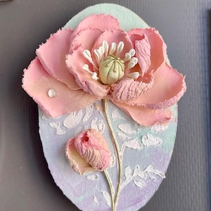 Peut inclure: Une œuvre d'art ovale faite à la main représentant une fleur rose aux pétales texturés et une tige dorée. La fleur est posée sur un fond pastel bleu et violet avec des accents blancs. Un petit bouton est également visible.