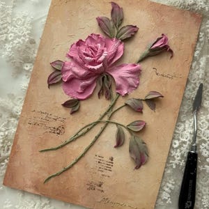 May include: A textured artwork featuring a pink rose with green stems and leaves, set against a beige background with script-like markings. A palette knife rests on the side. The artwork has a vintage, romantic aesthetic.