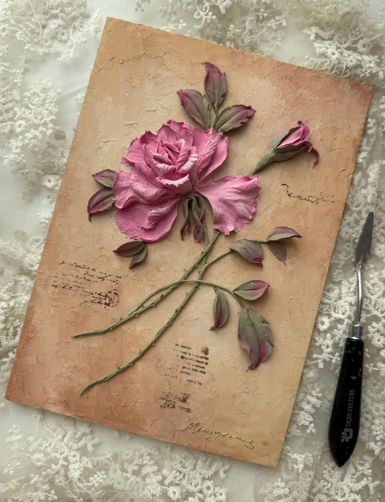 May include: A textured artwork featuring a pink rose with green stems and leaves, set against a beige background with script-like markings. A palette knife rests on the side. The artwork has a vintage, romantic aesthetic.