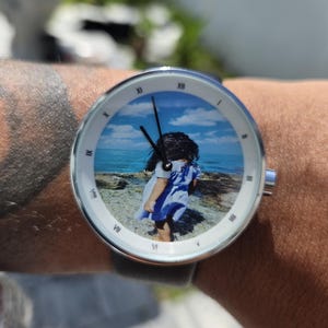 May include: A round wristwatch with a silver-toned metal case and a white bezel featuring black Roman numerals. The watch face displays a color photograph of a child standing on a rocky shore by the ocean under a blue sky with black hands.