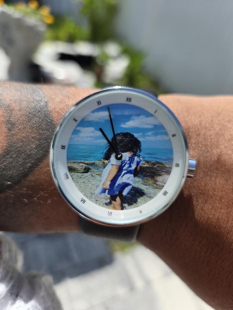 May include: A round wristwatch with a silver-toned metal case and a white bezel featuring black Roman numerals. The watch face displays a color photograph of a child standing on a rocky shore by the ocean under a blue sky with black hands.