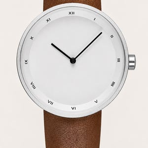 May include: A wristwatch with a silver-toned circular case and a plain white dial. The dial features black Roman numerals from I to XII around the inner edge. It has two thin black hands and a brown textured leather strap with a silver crown.