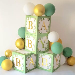 May include: A display of green blocks spelling "BABY", surrounded by white, gold, and green balloons. The blocks are light green with gold lettering and a leafy design. The balloons are in various sizes and colours.