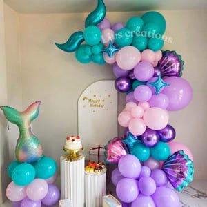 May include: A mermaid-themed birthday party display featuring a balloon arch in teal, purple, and pink. Includes a mermaid tail balloon, star-shaped balloons, and a white backdrop with "Happy Birthday" text. Cakes and gifts are also present.