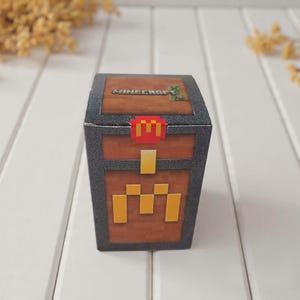 Puede incluir: Una caja con forma de cofre con temática de Minecraft. La caja presenta una textura de madera marrón con ribetes negros y detalles dorados. La parte superior de la caja tiene el logotipo de Minecraft y un personaje pixelado verde. Un logotipo rojo y amarillo está en el frente.