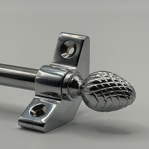 May include: A chrome curtain rod bracket with a decorative, textured finial. The bracket has a rectangular base with two mounting holes and a cylindrical rod holder. The finial is shaped like a pinecone.