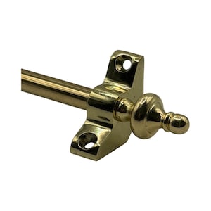 May include: A brass-coloured curtain rod with a decorative finial. The rod is attached to a bracket with two screw holes. The finial has a rounded shape with a small, pointed top. The hardware is made of metal.