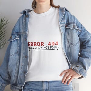 May include: A white t-shirt featuring the red and black text ERROR 404 MOTIVATION NOT FOUND, worn under a light blue denim jacket. The shirt has a crew neckline and short sleeves, while the denim jacket features metal buttons and chest pockets.