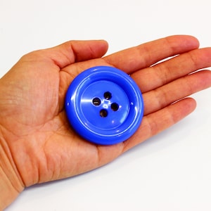 May include: A large, round, glossy blue button featuring a smooth, slightly raised outer rim and a recessed center area containing four evenly spaced holes for threading. The button has a vibrant, solid blue color and a polished, reflective finish.