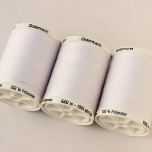 May include: Three white spools of Gutermann 100% polyester thread. Each spool is labeled with the Gutermann brand name and contains 1000 m - 1094 yds/vgs of thread. The spools are cylindrical with a white base.
