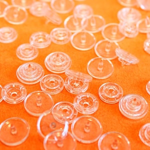 Puede incluir: Una colección de broches de presión de plástico transparente y circular dispersos por el encuadre. Cada broche presenta una superficie lisa y transparente con una punta central o una pequeña abertura circular para unir telas.