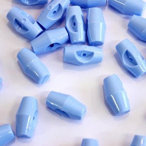 Pode incluir: Um conjunto de fechos de cordão de plástico azul claro. Cada fecho de cordão tem uma forma cilíndrica com um orifício central e dois orifícios laterais. Estão espalhados numa superfície branca.