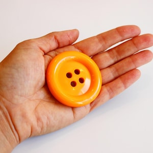 Può includere: Un grande bottone rotondo arancione brillante con quattro fori. Il bottone è tenuto in una mano aperta su uno sfondo bianco. Questo accessorio da cucito è un accento vibrante per abbigliamento o progetti artigianali.