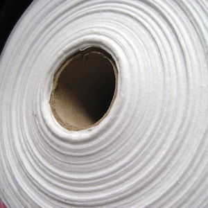 May include: Close-up view of a roll of white fabric. The fabric is tightly wound, showing concentric circles. The core is a brown cardboard tube. The fabric has a soft texture, with visible ridges and folds.