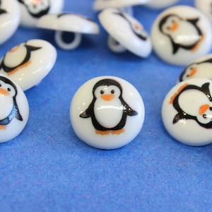 Op de afbeelding: Witte plastic knopen met pinguïnontwerpen. Elke knop heeft een zwart-witte pinguïn met een oranje snavel en poten.