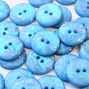 Puede incluir: Una colección de botones redondos de color azul claro con un acabado nacarado y jaspeado. Cada botón cuenta con dos orificios centrados para su fijación. La superficie de los botones tiene una textura lisa y brillante con variaciones.