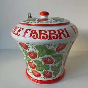 Può includere: Un vaso di ceramica bianco con coperchio, decorato con fragole rosse e foglie verdi. Il vaso ha la scritta "LE FABBRI" in rosso. Il coperchio ha un pomello rosso e un cucchiaio d'argento. La base e il bordo sono rossi.