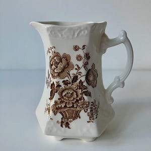 Op de afbeelding: Een crèmekleurige keramische kan met een bruin bloemenpatroon in transferdruk, met bloemen in een mand. De kan heeft een zeshoekige voet, een gebogen handvat en een gegoten decoratief detail langs de bovenrand van het vat.