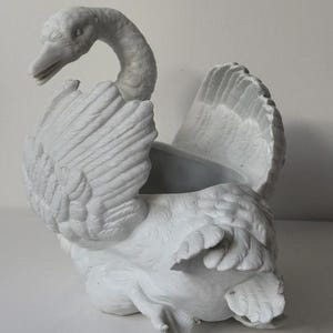 May include: A white ceramic swan-shaped planter featuring detailed moulded feathers on the wings and body. The swan has an arched neck and an open top cavity for holding items. The surface has a matte finish with visible sculpted textures.