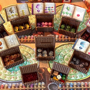 Puede incluir: Configuración de un juego de mesa con varios componentes. Varios cofres de madera marrón pequeños están abiertos, cada uno contiene piezas de juego de diferentes colores. Pequeños libros con ilustraciones y texto se colocan encima de los cofres. El tablero del juego presenta un diseño circular.