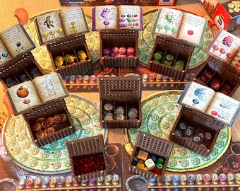 May include: A tabletop board game setup with various components. Several small, brown wooden chests are open, each containing different colored game pieces. Small books with illustrations and text are placed above the chests. The game board features a circular design.