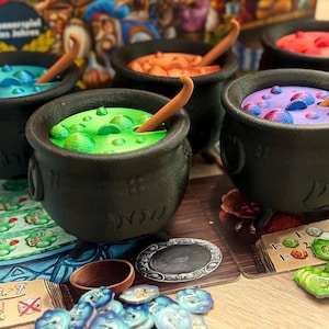 以下が含まれることがあります： カラフルな薬液が入った5つの黒い大釜が特徴のボードゲーム。各釜には青、緑、オレンジ、紫、赤など、異なる色の液体が入っており、木のスプーンが添えられています。ゲームの駒とカードが大釜の周りに散らばっています。