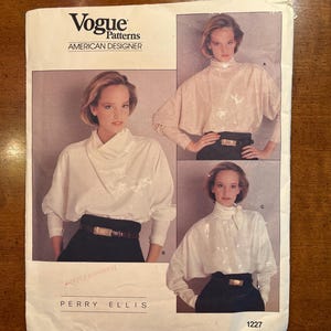 May include: A Vogue Patterns American Designer sewing pattern envelope for a Perry Ellis blouse, number 1227. The cover features three photographs of a white long-sleeved blouse with a high neckline, worn with a dark belt over dark trousers.