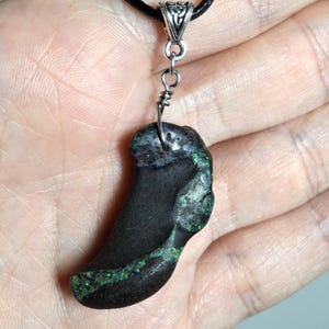 Puede incluir: Un collar con colgante negro con una piedra en forma de garra. La piedra tiene detalles iridiscentes verdes y azules. El colgante está sujeto a un cordón negro con un cierre de metal plateado.