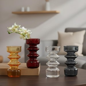 May include: Four glass vases in amber, red, clear, and gray. Each vase has a unique, stacked, rounded design. One vase holds white flowers. The vases are arranged on a dark wooden surface, with a neutral background.
