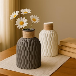 Op de afbeelding: Twee decoratieve vazen, één donkergrijs en één wit, met een textuur, golfachtig patroon. De vazen hebben een met touw omwikkelde hals en bevatten witte madeliefjes. De vazen staan op een neutraal gekleurde tafelloper.