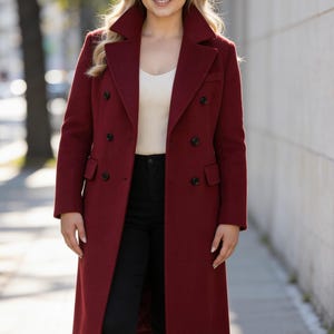 Puede incluir: Un abrigo largo burdeos de doble botonadura con botones negros. El abrigo tiene un cuello ancho y se lleva sobre una camiseta color crema y pantalones negros. La persona lleva botas negras. El fondo es una calle de la ciudad.
