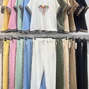 Può includere: Una collezione di completi loungewear a due pezzi abbinati in vari colori, tra cui giallo, rosa e blu. Ogni completo presenta un top a maniche corte con finiture leopardate e un paio di pantaloni abbinati con una striscia leopardata sul lato. Il top bianco ha un design a cuore colorato.