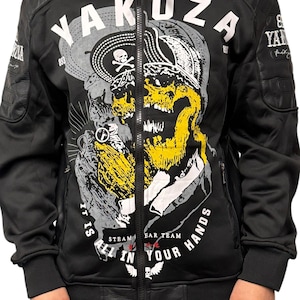 Puede incluir: Chaqueta negra con cremallera y un gráfico de calavera en amarillo, gris y blanco. La chaqueta presenta la palabra "Yakuza" en letras blancas grandes y el texto "IT IS ALL IN YOUR HANDS". La chaqueta tiene detalles de cuero en los hombros.
