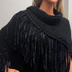 May include: A black knitted poncho with a high collar and fringe detail. The poncho is made of a thick, textured yarn and features long, dark fringe along the edges. The collar is a ribbed knit, and the overall design is simple yet stylish.