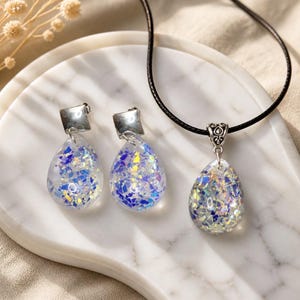 Peut inclure: Un ensemble de bijoux composé de deux boucles d'oreilles en forme de goutte et d'un pendentif assorti sur un cordon noir. Chaque pièce présente une base en résine transparente remplie de flocons irisés bleus et argentés, avec des détails en métal argenté.