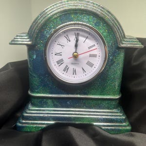 May include: A decorative quartz clock with a green and blue glitter finish. The clock has a white face with black Roman numerals and a red second hand. The clock is a small, tabletop size.