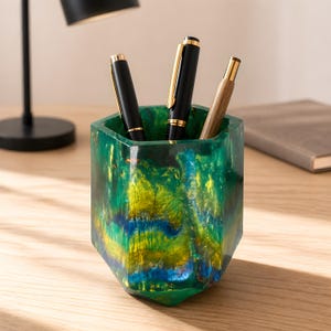 May include: A hexagonal pen holder made of resin with a swirling pattern of emerald green, bright yellow, and deep blue colours. The container holds two black pens with gold-tone clips and one light brown wooden pen with a gold-tone tip and cap.