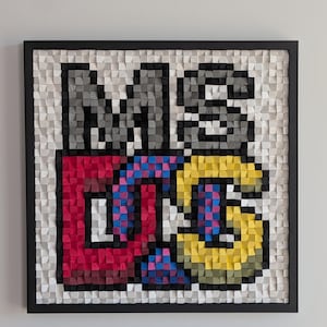 Op de afbeelding: Een vierkant wandkunstwerk met de tekst MS DOS, opgebouwd uit kleine, individuele houten blokjes. De letters zijn uitgevoerd in grijs, rood, blauw en geel tegen een witte achtergrond, alles gevat in een dunne zwarte lijst.